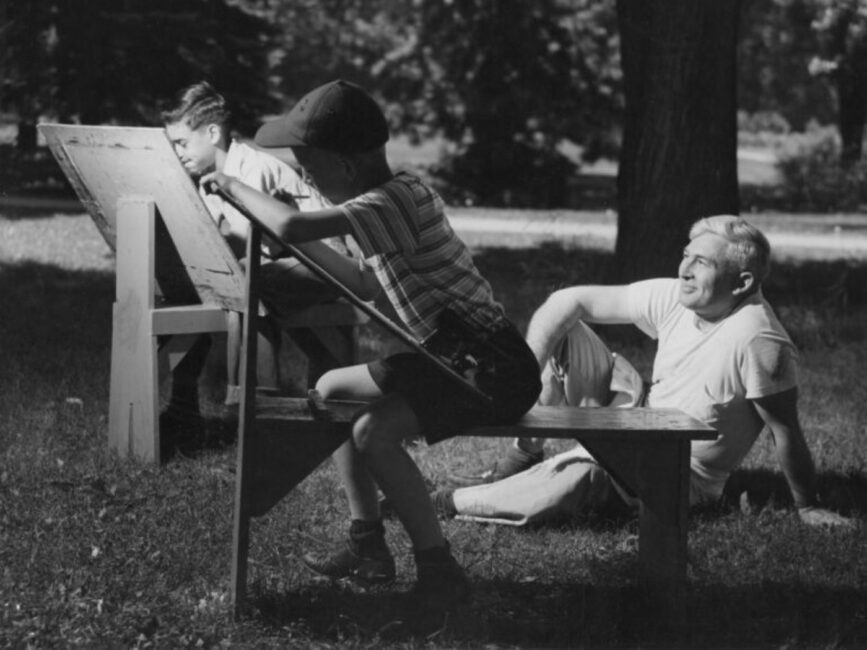 A black and white photo of two boys painting outside alongside an older man