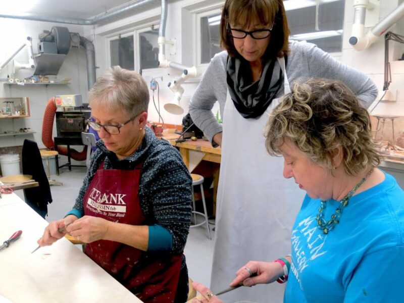 two adult students working on jewelry objects with an instructor overlooking them